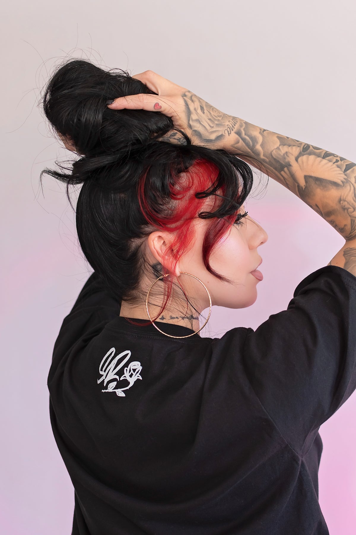 Person with black and red hair, wearing a black sweatshirt with a white logo, against a light pink background.