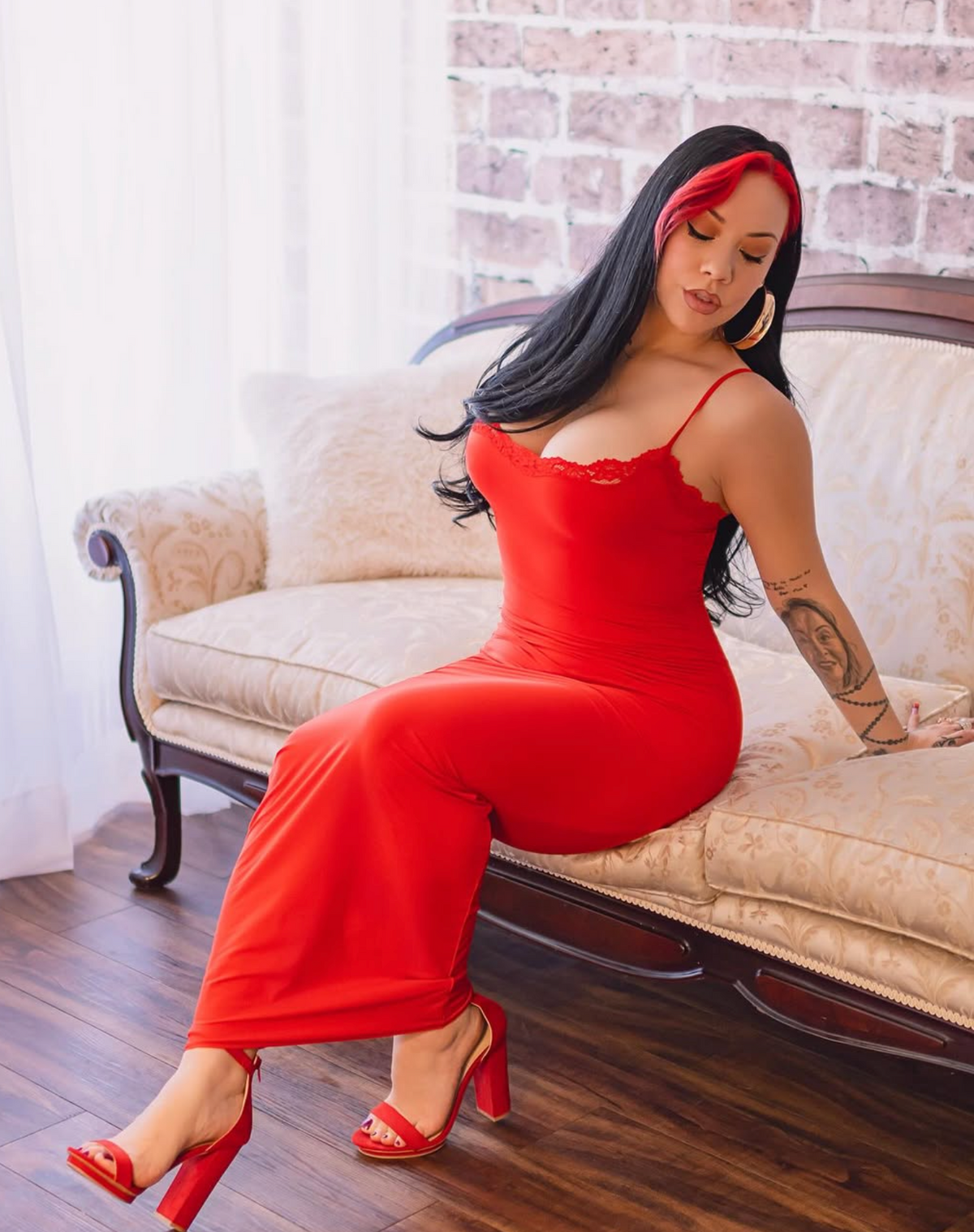 A woman wearing a bright red bodycon dress and matching red high heels sits on a beige patterned sofa in a living room with a brick wall and wood floor.4