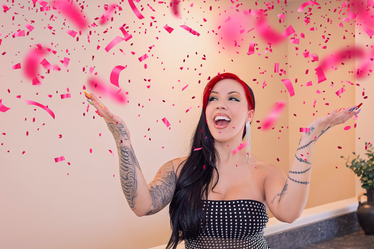 A woman with long black hair with a red money piece stands indoors with her arms raised, surrounded by falling pink confetti. She is wearing a strapless black and white top.